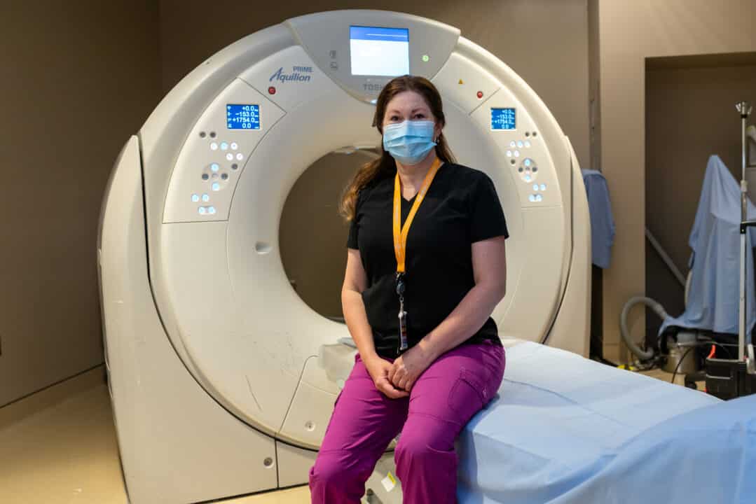 A technologist sits on the table of a CT scanner.