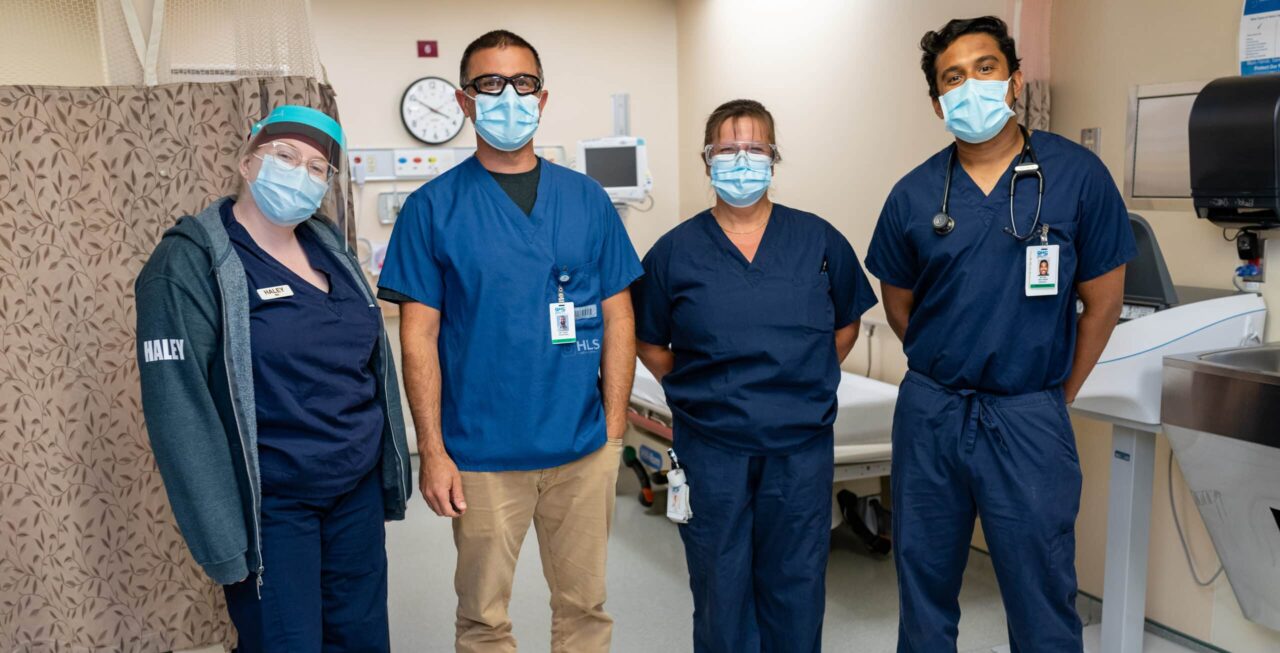 Four health care professionals in blue scrubs stand together in an emergency department.