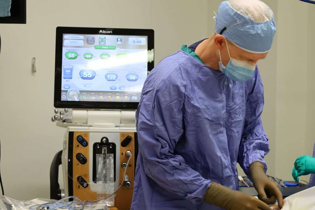 A surgeon wearing scrubs and a cap prepares for surgery. A monitor with the patient’s vitals is behind him.