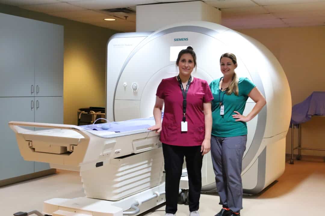 Two technologists stand beside an MRI machine.