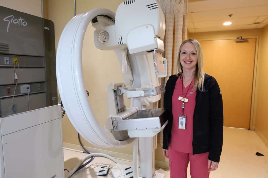 A smiling technologist stands with a mammography machine.