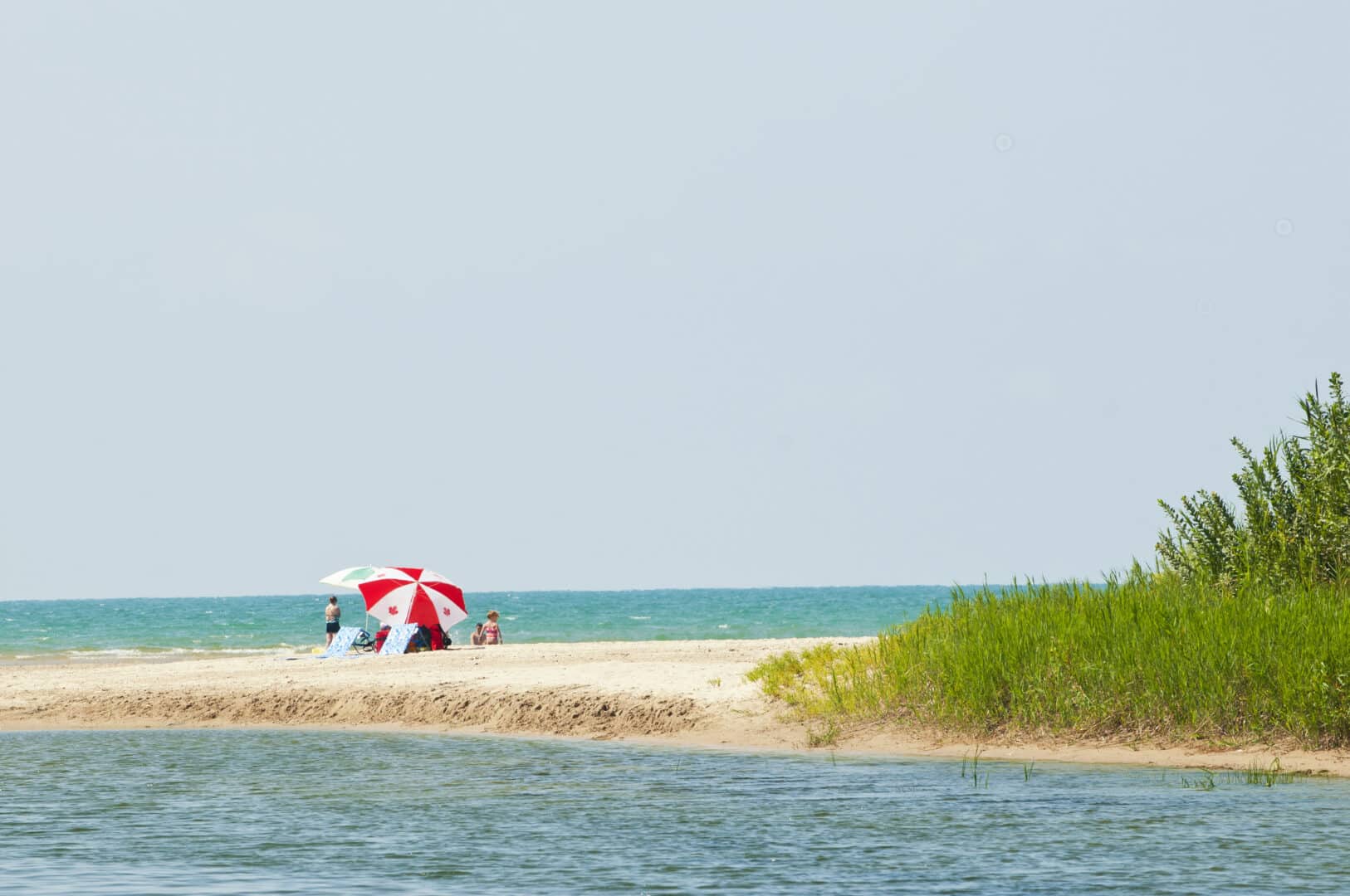 A family on a narrow, sandy beach with a Canada flag umbrella set up and beautiful blue water surrounding the sandy beach on both sides.