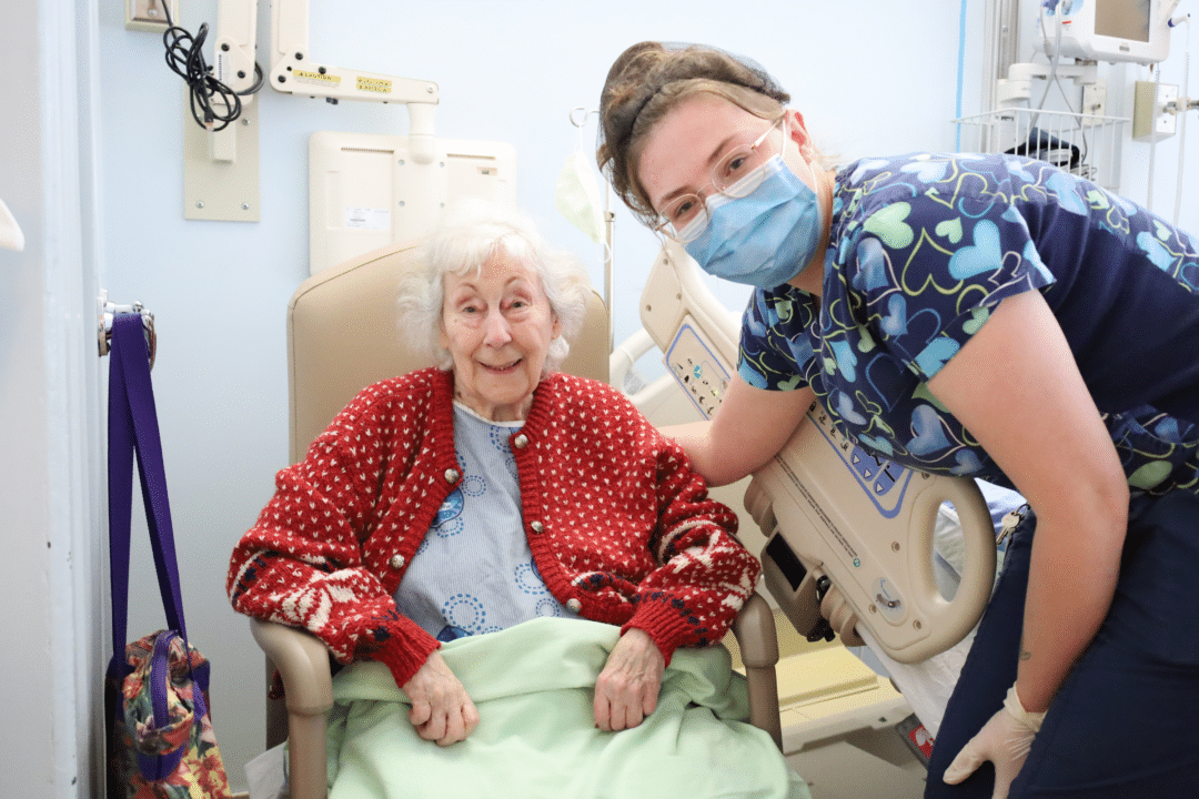 A hospital worker wearing a mask poses with an elderly patient who is smiling.