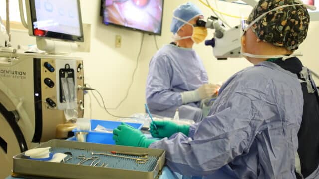 A surgeon looks through a magnifying eye piece while a surgical nurse looks at him and holds a surgical instrument.