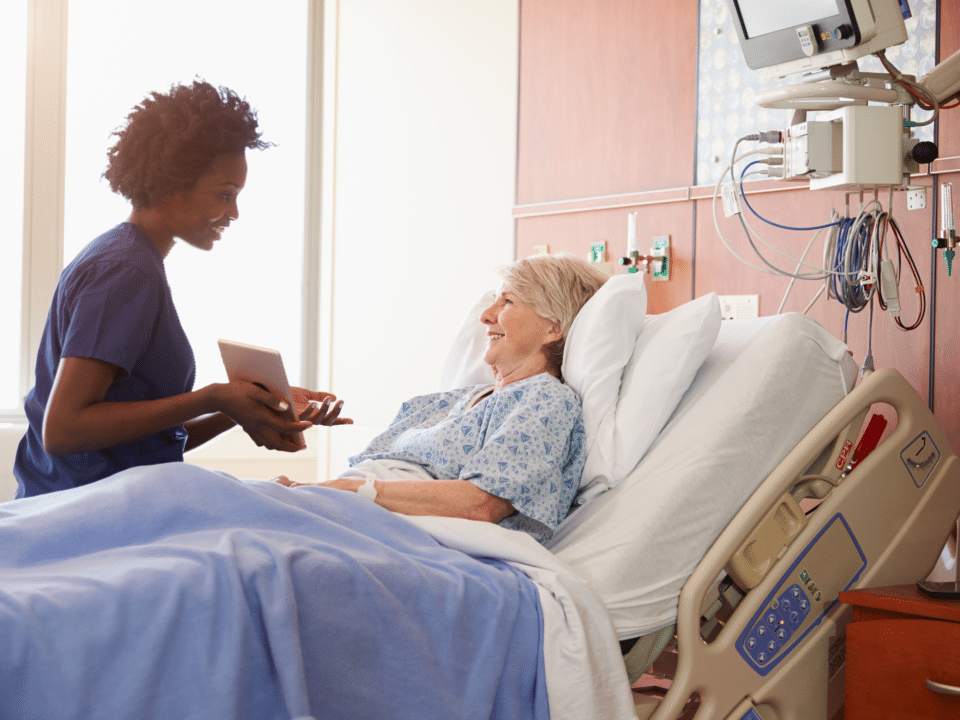 Caregiver sitting at the hospital bedside showing elderly patient a tablet.
