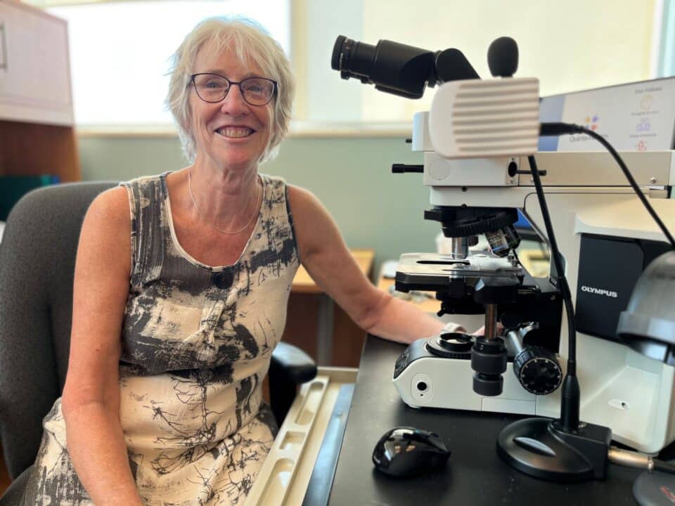 A pathologist sits in front of her microscope and smiles at the camera.