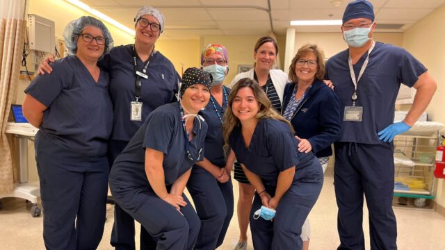 A group of eight smiling hospital workers gather together in a surgery recovery room.