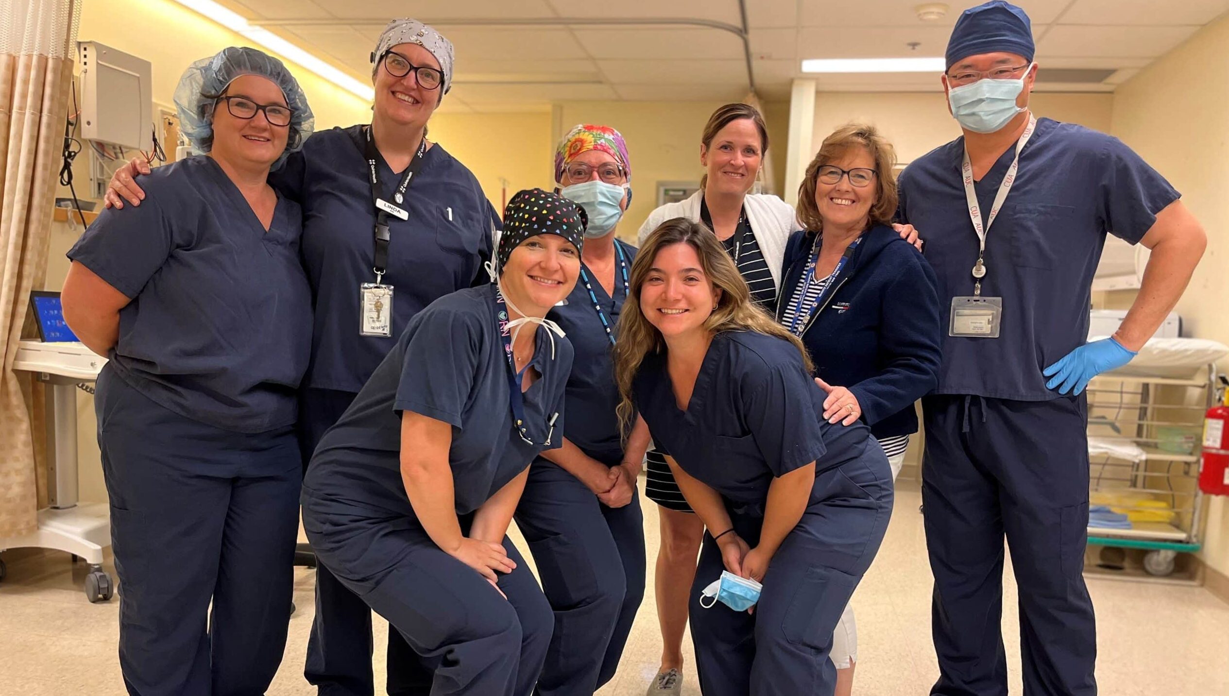 A group of eight smiling hospital workers gather together in a surgery recovery room.