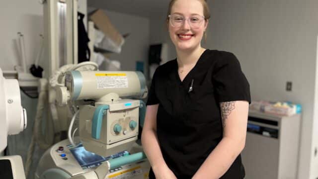 An X-ray tech stands in front of imaging equipment and smiles.