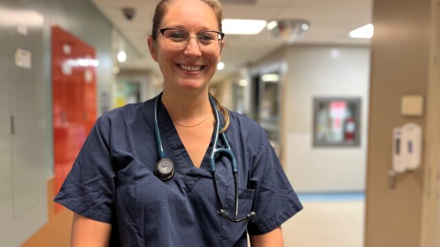 A doctor wearing scrubs with a stethoscope around her neck stands in an emergency department.