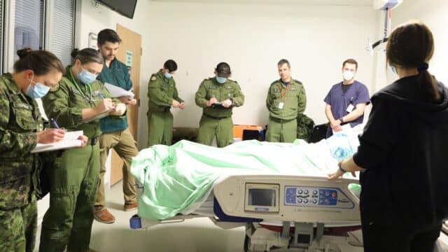 Military and hospital staff gather around a hospital bed and take notes.