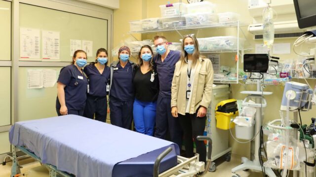 A group of six health care workers gather in the emergency department trauma room.