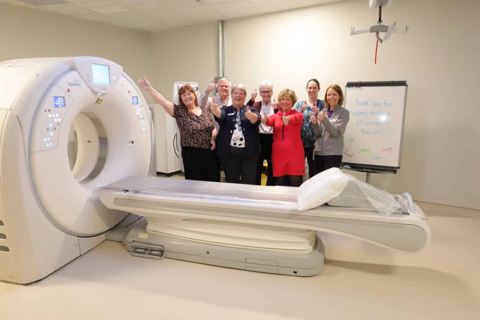 Seven people stand beside a CT machine giving thumbs up.