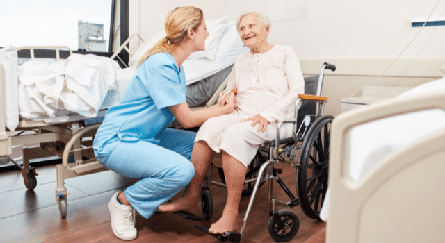 A healthcare worker squats and smiles with an elderly patient in a wheelchair.