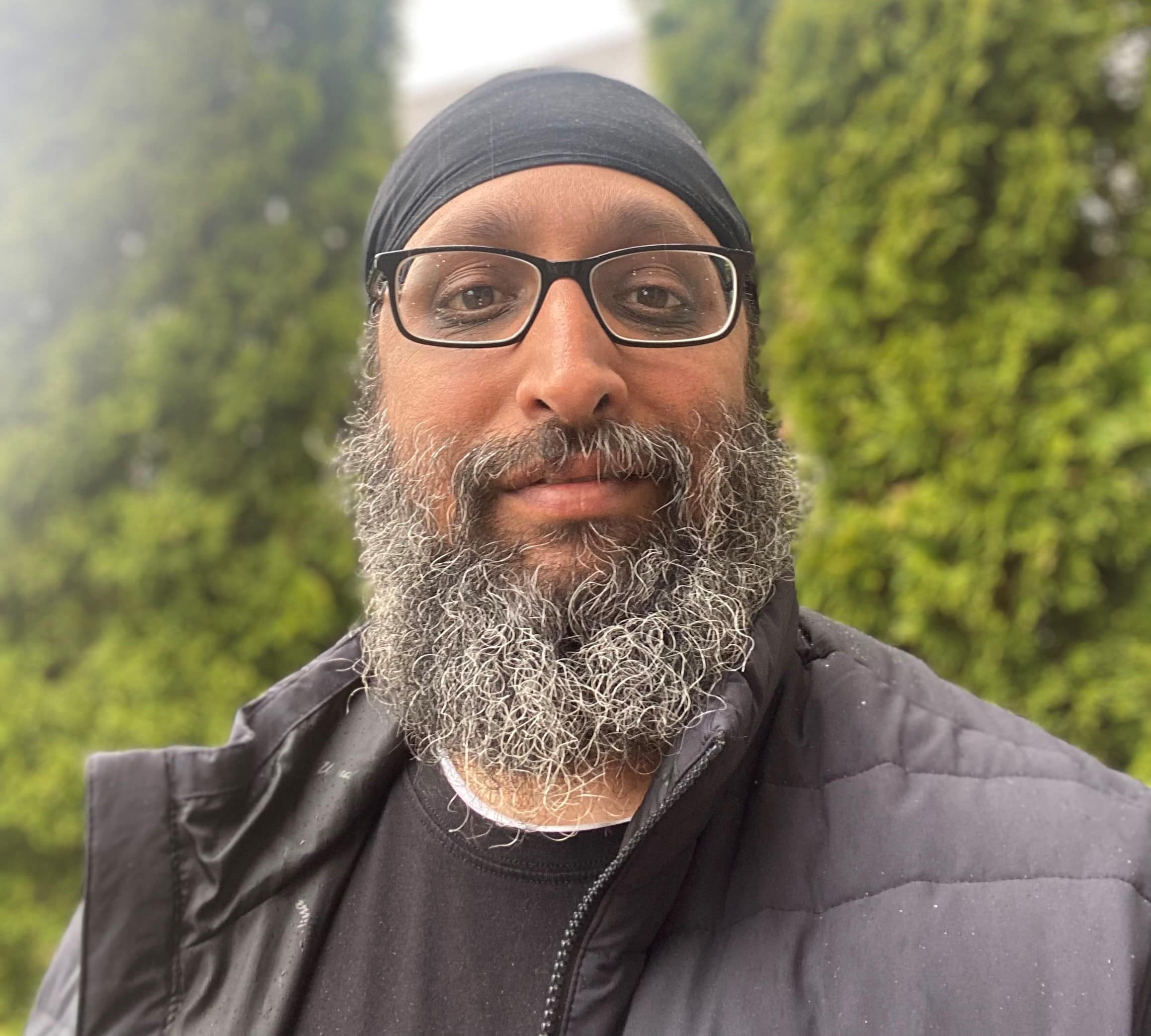 A man with a beard, head covering and glasses standing outside in front of a bush.