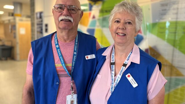 An older couple wearing blue vests and lanyards smile in a lobby.