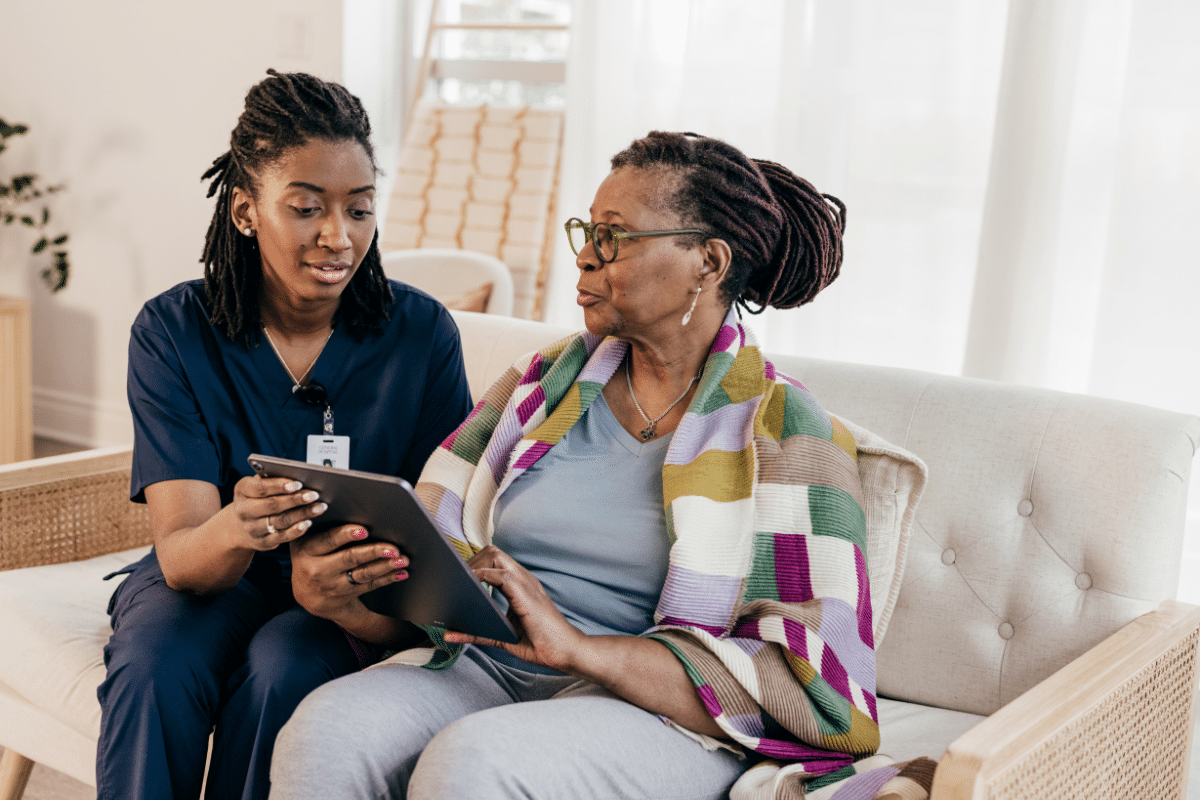 A younger woman wearing scrubs and a badge sits on a couch with an older woman and they look at an iPad together.