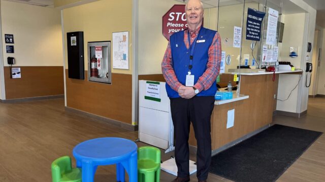 An older man with white hair wearing a blue vest stands beside a plastic children's table and two chairs in the emergency department.