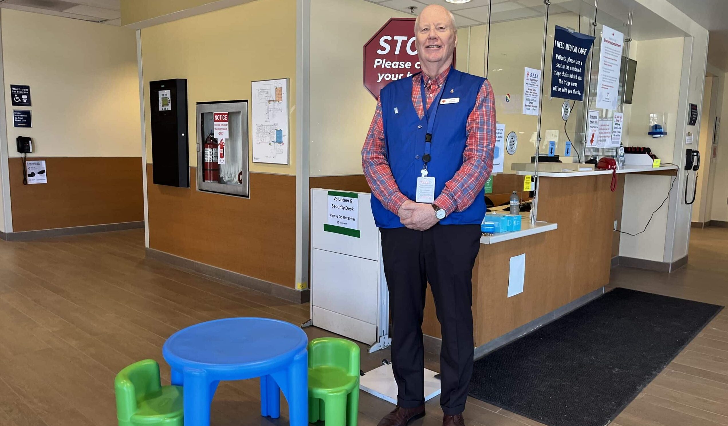 An older man with white hair wearing a blue vest stands beside a plastic children's table and two chairs in the emergency department.