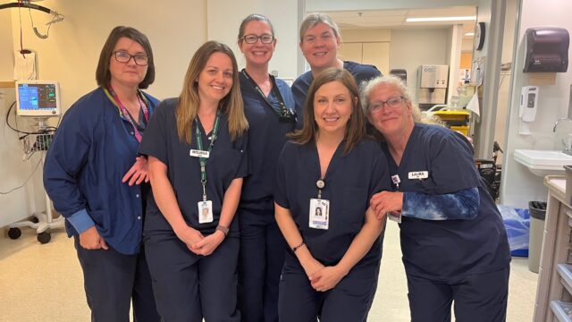 Six women wearing blue scrubs gather together in a surgical area.