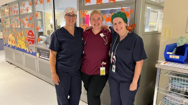 Three female health care professionals wearing scrubs stand together on a maternity unit.